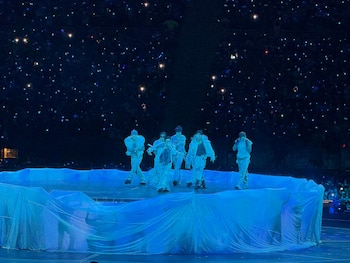 Integrantes de un grupo musical cantan sobre un escenario circular iluminado de azul ante un público a oscuras con pequeñas luces