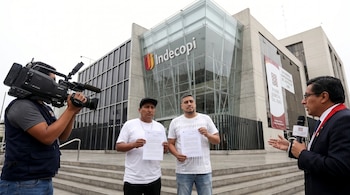 Un camarógrafo filma a dos hombres que sostienen documentos frente al edificio de Indecopi, mientras un hombre con micrófono del Poder Judicial habla a la derecha.