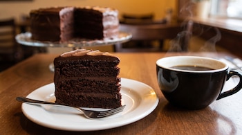Porción de torta de chocolate en un plato blanco con tenedor, una taza de café negro humeante y la torta completa de fondo.