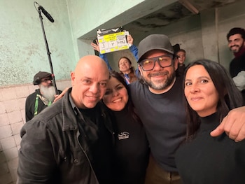 Um grupo de seis pessoas sorridentes em um quadro de filme, com um microfone à esquerda e um quadro amarelo e preto ao fundo.