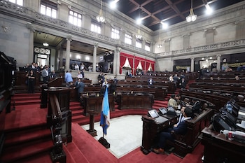 Interior de un hemiciclo parlamentario con filas de escaños de madera, alfombra roja y varias personas. Se aprecia la bandera de Guatemala al frente