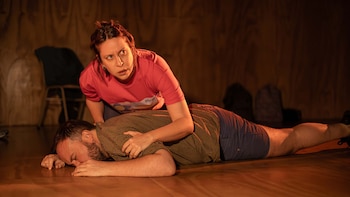 Imagen de una actriz arrodillada junto a un actor acostado en el suelo de un escenario teatral, la mujer mira hacia arriba con preocupación