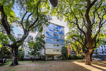 Vista de un edificio con un mural que simula el cielo con nubes, flanqueado por grandes árboles de frondosas copas verdes y un terreno de tierra y pasto