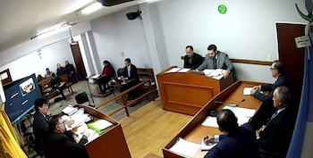Vista de una sala de audiencias judicial con personas en mesas y bancos, documentos sobre los escritorios, una pantalla y una bandera