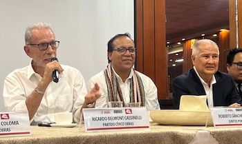 Tres hombres sentados a una mesa. Uno sostiene un micrófono. En la mesa hay placas identificativas con nombres, logos, vasos y una taza