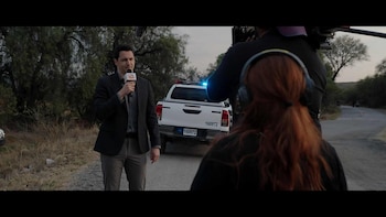 Carlos Gatica, vestido de traje, sostiene un micrófono frente a una cámara. Detrás, una camioneta blanca con luces policiales encendidas en una carretera rural