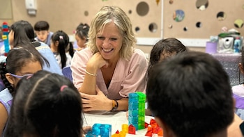 Una mujer rubia con blusa rosa claro sonríe sentada a una mesa con varios niños, construyendo con bloques translúcidos de colores