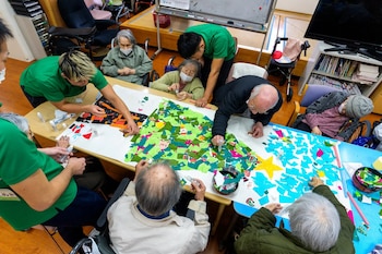 Ancianos y personal pegan trozos de papel de colores sobre una mesa para crear un mural navideño con un árbol verde y un cielo azul con estrellas