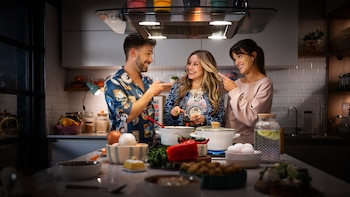 Karol Sevilla, sonriente, en una cocina moderna con dos acompañantes. Los tres interactúan alrededor de una isla con ollas e ingredientes para cocinar