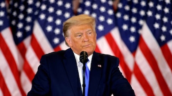 El Presidente de EE. UU. Donald Trump, con traje azul y corbata, habla en un micrófono. Detrás de él, varias banderas de Estados Unidos