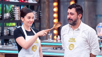 Una mujer y un hombre usan delantales blancos con el logo de MasterChef. La mujer sostiene una cuchara de madera, el hombre tiene barba y expresión triste.