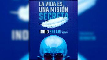 Portada del libro 'La vida es una misión secreta' de Indio Solari sobre fondo azul, con una pluma blanca y la parte superior de una cabeza con gafas de sol