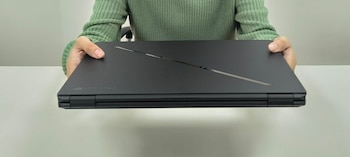 Manos de una persona con suéter verde sostienen un portátil negro cerrado con una línea diagonal en la tapa sobre una superficie clara