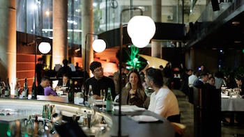 Varias personas conversan sentadas en la barra curva de un restaurante iluminado con lámparas de globo