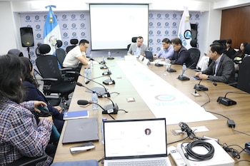 Sala de conferencias con once personas, mesa larga, micrófonos, ordenadores portátiles, botellas de agua, proyector, dos banderas y logos VALOR
