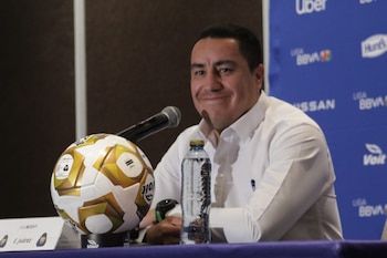Hombre en camisa blanca sonriendo en un podio con micrófono, balón de fútbol blanco y dorado, y botella de agua, frente a un fondo azul con logos de patrocinadores