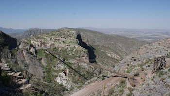 Paisaje montañoso con ruinas de edificios de piedra, un puente metálico largo, un caballete de madera y vegetación escasa bajo un cielo azul despejado