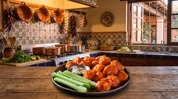 Plato de boneless de pollo picantes, apio fresco y aderezo sobre una mesa de madera. Al fondo, una cocina mexicana con azulejos decorados y ollas de cobre.