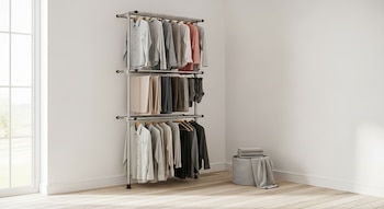 Tendedero vertical de metal con tres niveles de ropa colgada, incluyendo camisas, pantalones y calcetines, en un pasillo con paredes blancas y suelo de madera clara. Una canasta de ropa gris con toallas dobladas está a la derecha.