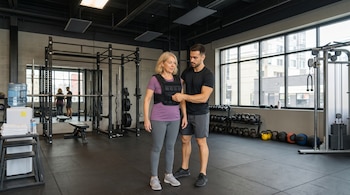 Mujer rubia con chaleco de peso y hombre en gimnasio. El lugar tiene estantes, pesas rusas, mancuernas, máquina de cables y ventanales.