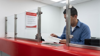 Un hombre con camisa azul sostiene un cheque detrás de un panel de vidrio sobre un mostrador rojo en una oficina.