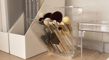 Pinceles de maquillaje surtidos en un organizador de acrílico transparente sobre una mesa de madera clara, con cajas de almacenamiento blancas en el fondo.
