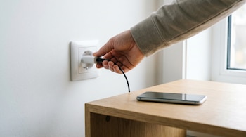 Una mano conectando un cargador USB en un enchufe de pared blanco; un teléfono móvil negro yace sobre una mesa de madera clara a la derecha.