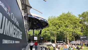 Cartel con "Capital One CityParks SummerStage" a la izquierda, un escenario al aire libre y personas sentadas en el césped. Se ven árboles verdes al fondo