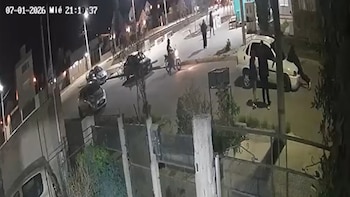 Edificios, postes de luz, calle asfaltada, dos automóviles oscuros estacionados, un automóvil blanco, una motocicleta, varias personas. Fecha y hora en pantalla