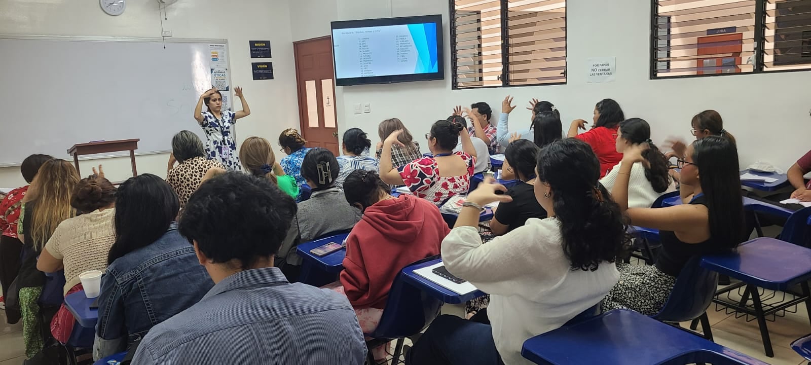 Una ponente expone ante un grupo de asistentes que levantan las manos durante un seminario de capacitación en un aula.