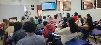 Personas sentadas en un aula gesticulan con las manos frente a una expositora, una pantalla y un pizarrón