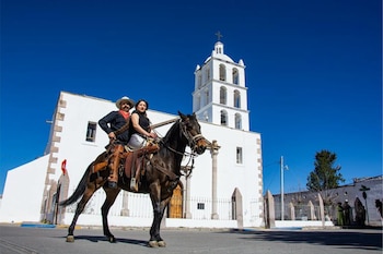 Hombre con sombrero y mujer montan un caballo oscuro frente a una iglesia blanca con campanario de arcos bajo un cielo azul