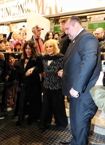 Mirtha Legrand, acompañada por una mujer y un guardia de seguridad, camina entre una multitud con teléfonos cerca de la entrada de un teatro con cartel