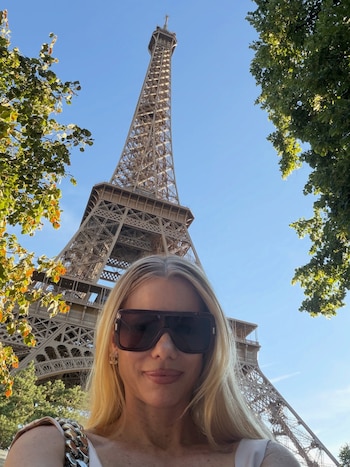 Una mujer rubia con gafas de sol oscuras se toma una selfie con la Torre Eiffel al fondo, rodeada de árboles y cielo azul