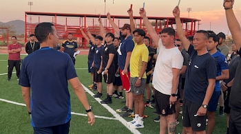 Varios hombres alineados en una cancha de césped artificial levantan tarjetas de colores ante un instructor en primer plano.
