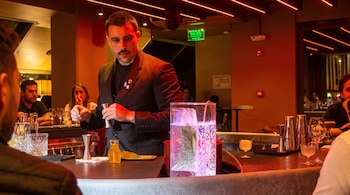 Un hombre con bigote detrás de la barra de un bar con varias copas, medidores, cocteleras y un recipiente alto con líquido y hierbas.