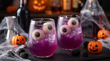 Dos vasos con bebida morada, hielo, zarzamoras y lichis con moras azules, acompañados por pequeñas calabazas y telarañas.