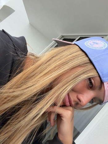 Primer plano de una mujer con cabello rubio largo que usa una gorra celeste y rosa, con la mano izquierda apoyada en la barbilla y fondo claro