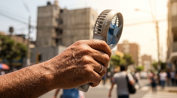Una mano con gotas de sudor sostiene un pequeño ventilador portátil en una calle urbana con edificios al fondo.