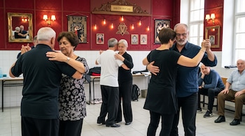 Tres parejas de adultos bailan abrazadas en el centro de una sala con pared roja, mientras otros dos hombres observan sentados.