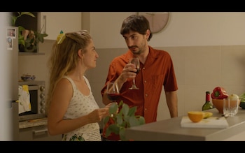 Hombre y mujer en una cocina, ambos sosteniendo copas de vino tinto. La mujer tiene un broche de limón en el cabello. Se ven ingredientes como pimiento rojo y naranja