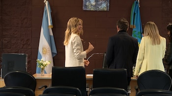 Una mujer de traje claro sonríe frente a tres personas de espaldas en una sala con la bandera argentina y sillas en primer plano.