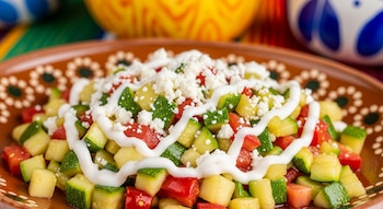 Plato de barro artesanal con calabacitas, jitomate y chile en cuadritos, cubierto con queso fresco desmoronado y un hilo de crema, sobre un fondo mexicano.