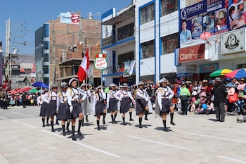 Grupo de estudiantes uniformados marcha en desfile cívico por una calle urbana. Hay una bandera de Perú, edificios y espectadores