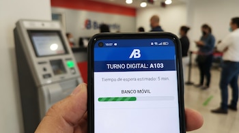 Una mano sostiene un smartphone mostrando la pantalla de una aplicación bancaria con un turno digital, un cajero automático y clientes borrosos en el fondo.