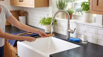 Unas manos sostienen una cuchara de madera bajo el agua de un grifo sobre un fregadero blanco en una cocina con azulejos.