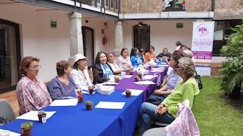 Un grupo de mujeres permanece sentado frente a mesas largas en un patio interior junto a pequeñas macetas y un cartel informativo