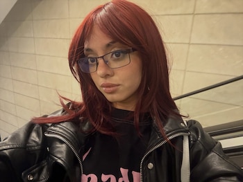 Joven con cabello rojizo, flequillo, gafas, chaqueta de cuero negra y camiseta negra. Fondo de pared de azulejos claros
