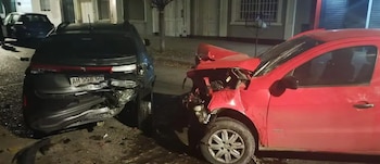 Vista nocturna de un choque entre dos autos en una calle urbana; un coche rojo con el capó abollado y uno gris con la parte trasera destrozada