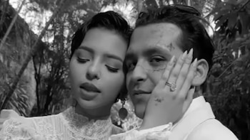 Primer plano en blanco y negro de Christian Nodal con tatuajes faciales, y Ángela Aguilar en un vestido de encaje, con su mano anillada sobre la mejilla de Nodal